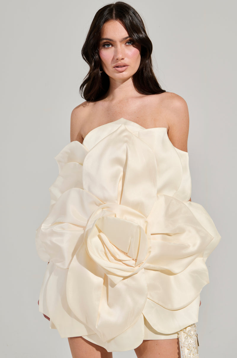 A woman with long dark hair models the FLOWER POWER MINI DRESS, a strapless design featuring an oversized 3D flower on a satin base, set against a light-colored plain background. 1