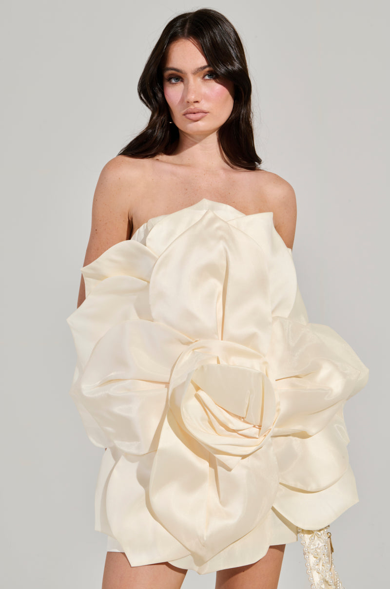 A woman with long dark hair is wearing the elegant FLOWER POWER MINI DRESS, which features an off-the-shoulder design crafted from a satin base and adorned with large, cream-colored floral fabric shapes. The 3D flower design of this dress creates a dramatic and elegant look as she stands against a plain background. 2