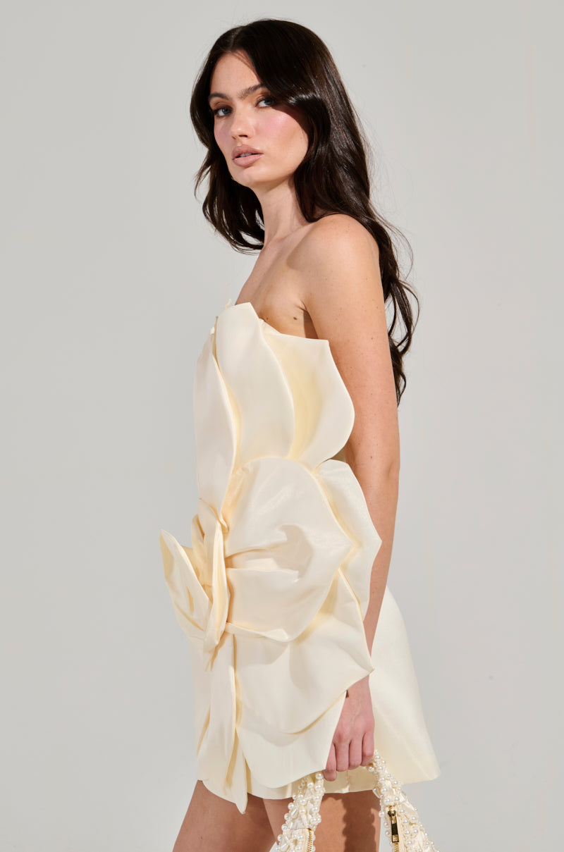 A model showcases the strapless FLOWER POWER MINI DRESS, adorned with a 3D flower pattern featuring large cream-colored fabric petals on a satin base, set against a simple background. She carries a textured light-colored purse while her long, dark hair cascades over her shoulders. 3