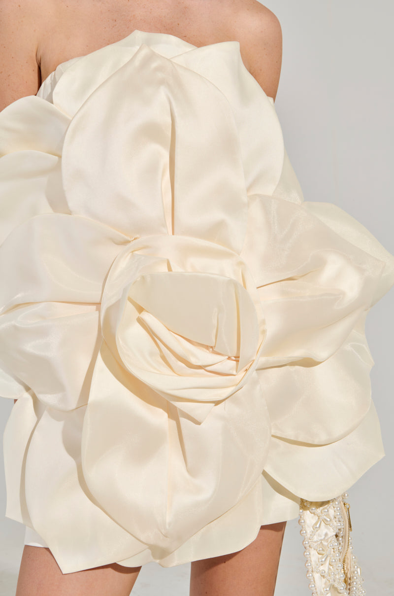 A woman models the FLOWER POWER MINI DRESS, a strapless garment featuring an intricate 3D floral pattern in a soft beige hue on a satin base, as she stands against a neutral backdrop with her right arm resting by her side. 7