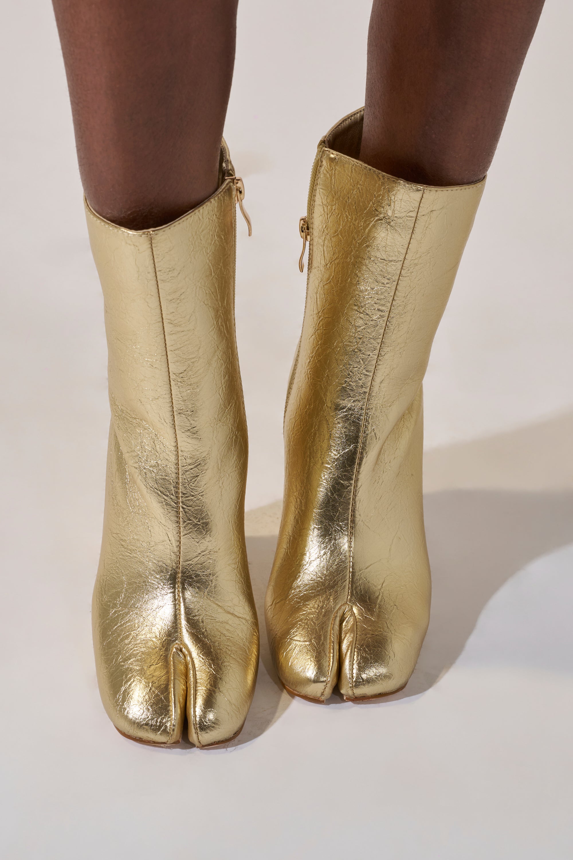 A close-up of someone wearing the AZALEA WANG BILHA GOLD SPLIT TOE CHUNKY BOOTIE, featuring shiny gold material and a distinctive split-toe ("tabi") design, as they stand on a light surface.