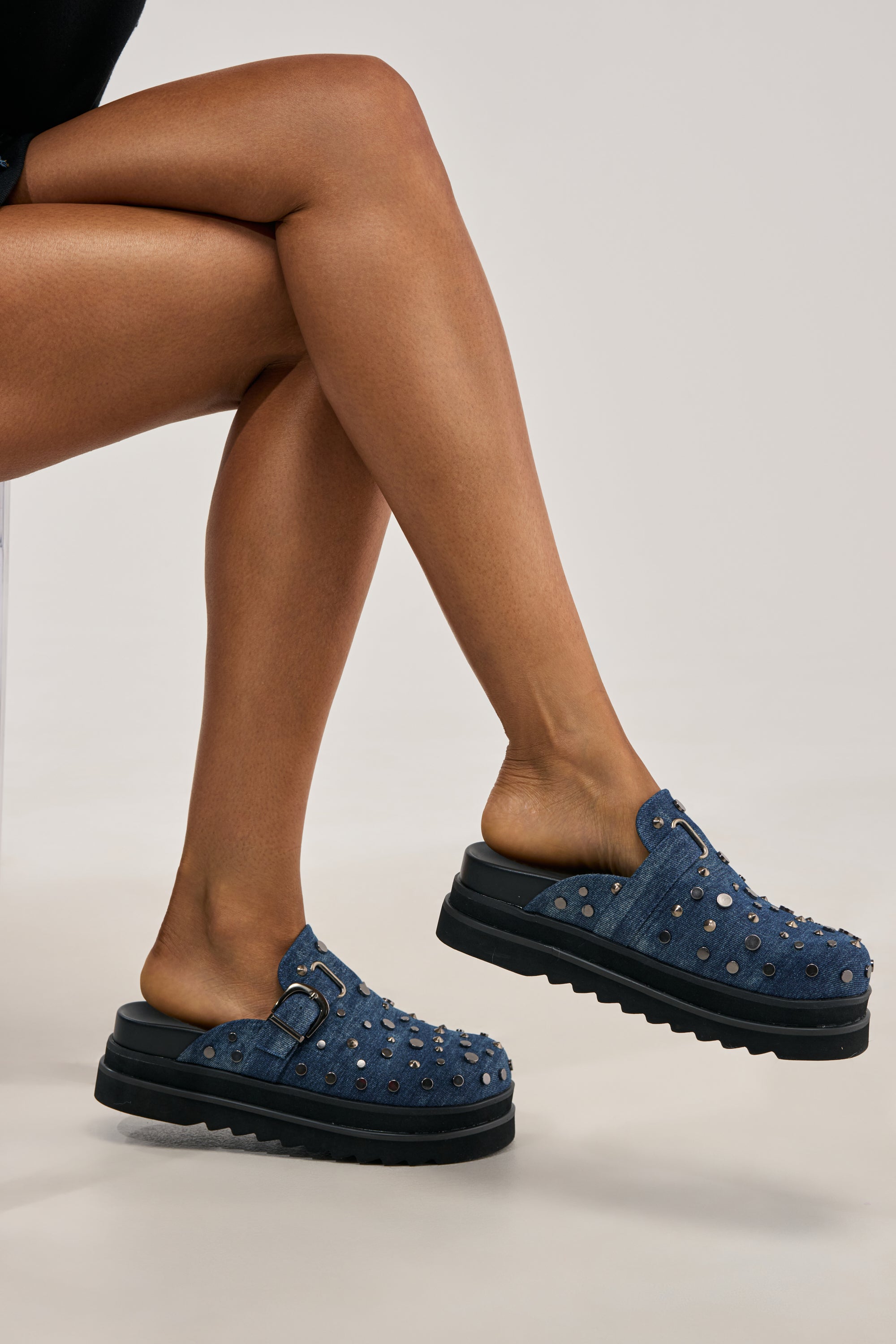 A person sits with legs crossed, showcasing the AKIRA GOLD BILLIANNA DENIM FLAT CLOG WITH HARDWARE—chic summer footwear featuring blue denim, platform soles, metallic studs, and buckle detailing—against a plain light background.