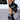 A person showcases the AZALEA WANG BIRD SONG black snap detail open toe bootie, featuring artistic leather wraps and metallic studs, paired with a matching black handbag accented with metal. The minimalist scene highlights these fashion details.