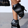 A person showcases the AZALEA WANG BIRD SONG black snap detail open toe bootie, featuring artistic leather wraps and metallic studs, paired with a matching black handbag accented with metal. The minimalist scene highlights these fashion details.