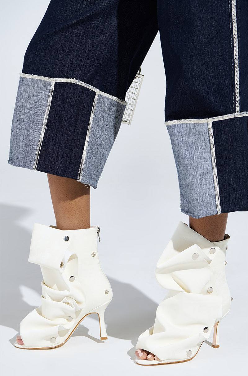 A person is wearing stylish dark blue and gray frayed denim pants paired with the unique AZALEA WANG BIRD SONG SNAP OPEN TOE BOOTIE IN BONE. The photo focuses on the lower legs and footwear against a light background.