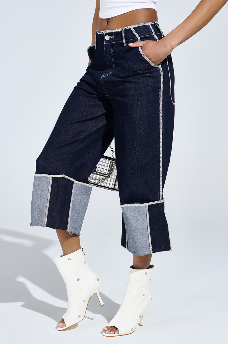 Model showcasing two-tone denim culottes with large cuffs and AZALEA WANG BIRD SONG SNAP OPEN TOE BOOTIE IN BONE against a plain background. The jeans flaunt visible white stitching and a raw hem, while the pose highlights the outfit's style and fit.