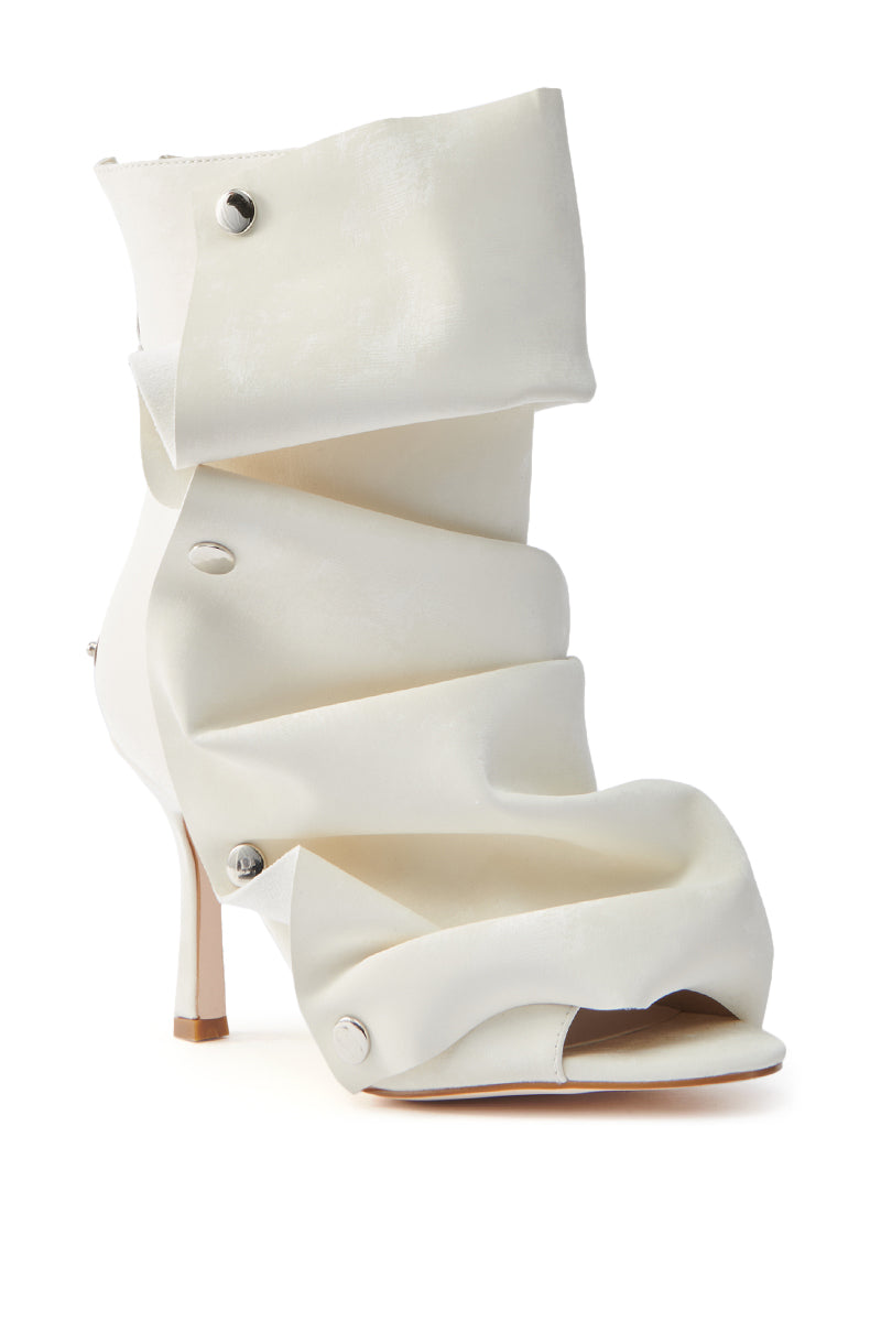 The AZALEA WANG BIRD SONG SNAP OPEN TOE BOOTIE IN BONE is a unique high-heeled shoe crafted from white fabric, featuring a spiral design with metallic button accents. Its narrow stiletto heel and peep-toe style create an artistic and elegant look against the white background.