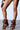The AZALEA WANG BIRD SONG BROWN SNAP DETAIL OPEN TOE BOOTIE, with distinctive high heels and button features, is worn with a small round brown leather handbag against a plain white wall.