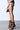 A person in profile wears a floral top, beige shorts, and AZALEA WANG BIRD SONG brown snap detail open-toe booties. They hold a small brown textured handbag against a plain white and gray wall and floor backdrop.
