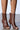 Close-up of legs in AZALEA WANG BIRD SONG brown snap detail open-toe booties. These faux leather booties feature a dark brown hue, small silver button accents, and a chic stiletto heel against a plain light-colored background.