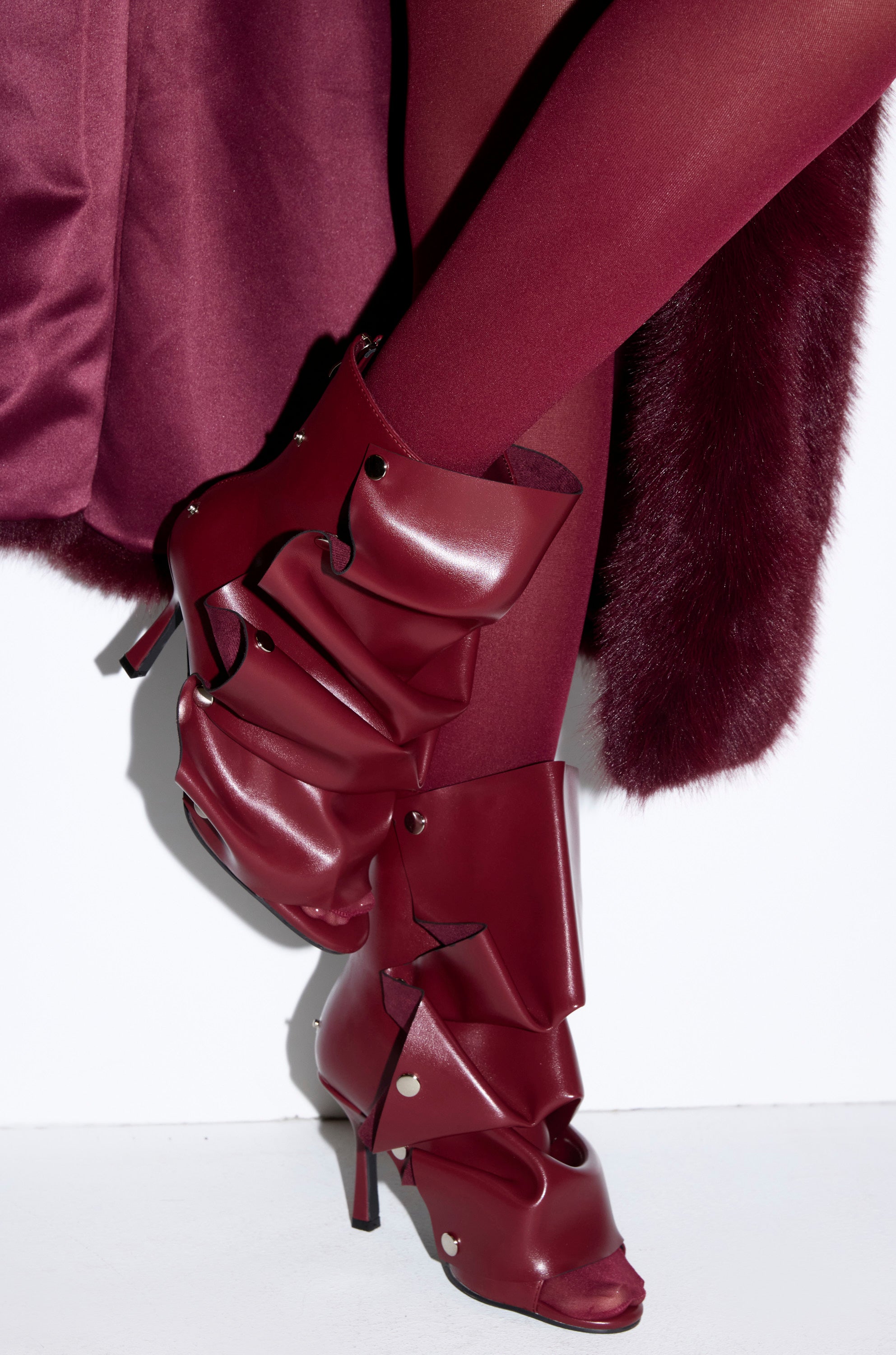 A person wearing burgundy tights, a matching skirt, and AZALEA WANG BIRDSONG BURGUNDY SNAP OPEN TOE STILETTO BOOTIES with layered, ruffled leather stands against a white background.