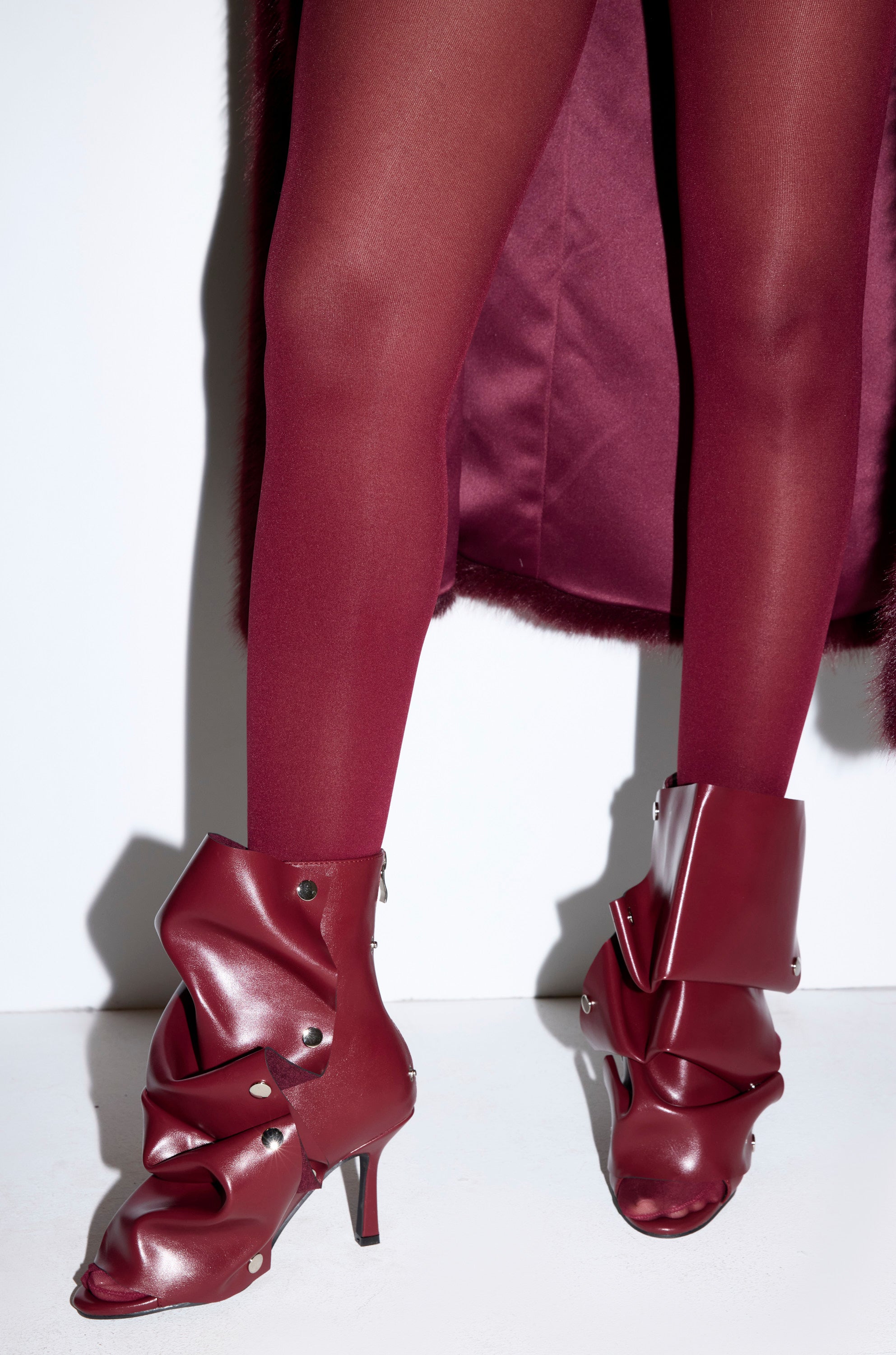 A person wearing sheer burgundy tights and AZALEA WANG BIRDSONG BURGUNDY SNAP OPEN TOE STILETTO BOOTIE stands against a white background.