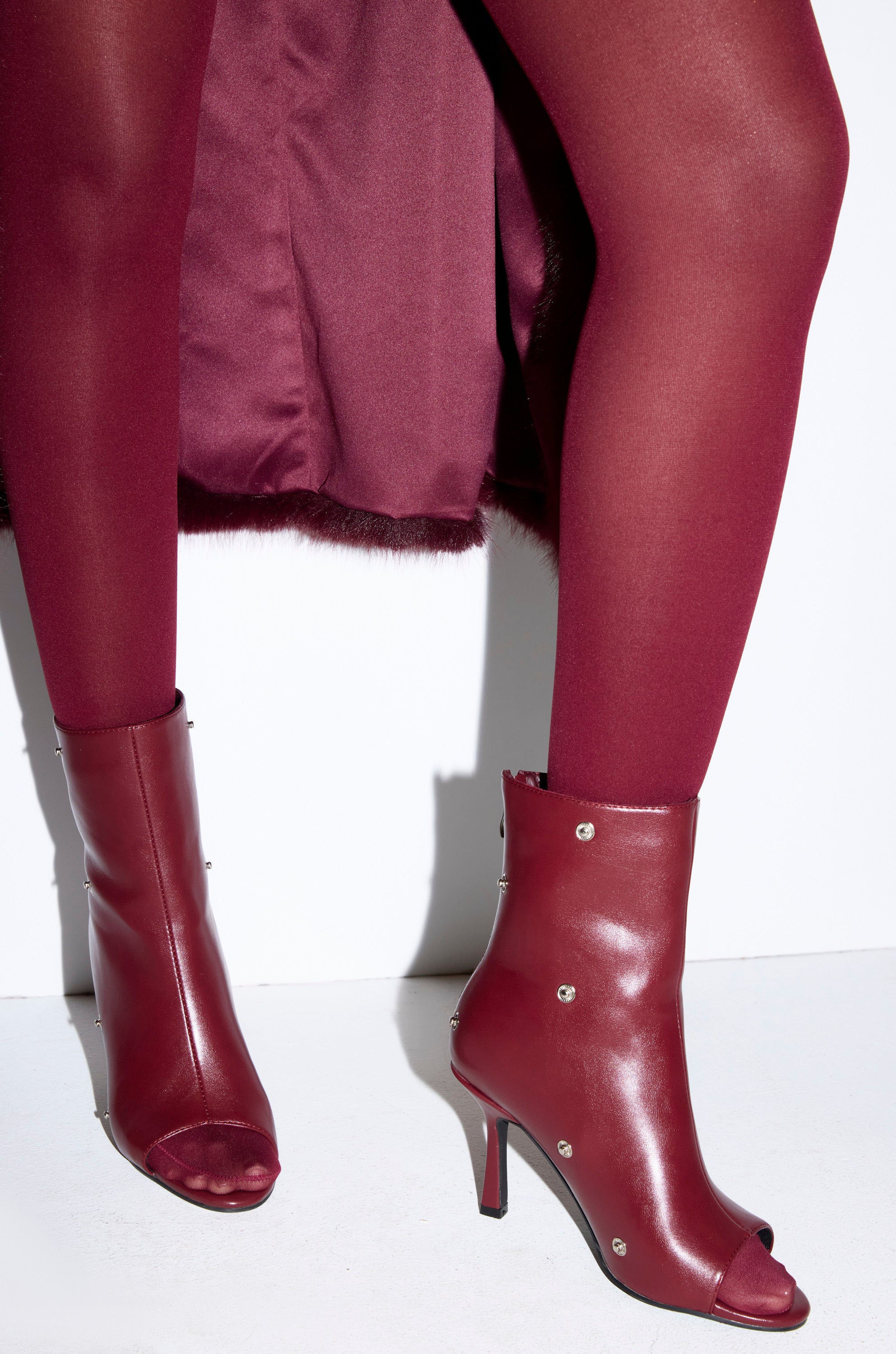 A person wearing burgundy tights, an elegant burgundy coat, and AZALEA WANG BIRDSONG BURGUNDY SNAP OPEN TOE STILETTO BOOTIE heels with metallic studs, standing against a white background.