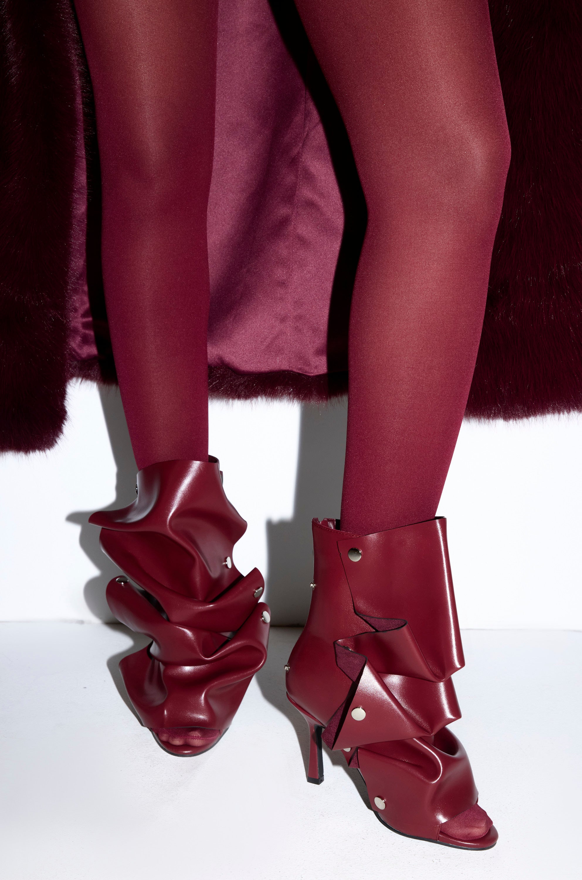A model in burgundy tights and a matching coat stands against a white background, showcasing the AZALEA WANG BIRDSONG Burgundy Snap Open Toe Stiletto Bootie with ruched, shiny leather and metallic studs.