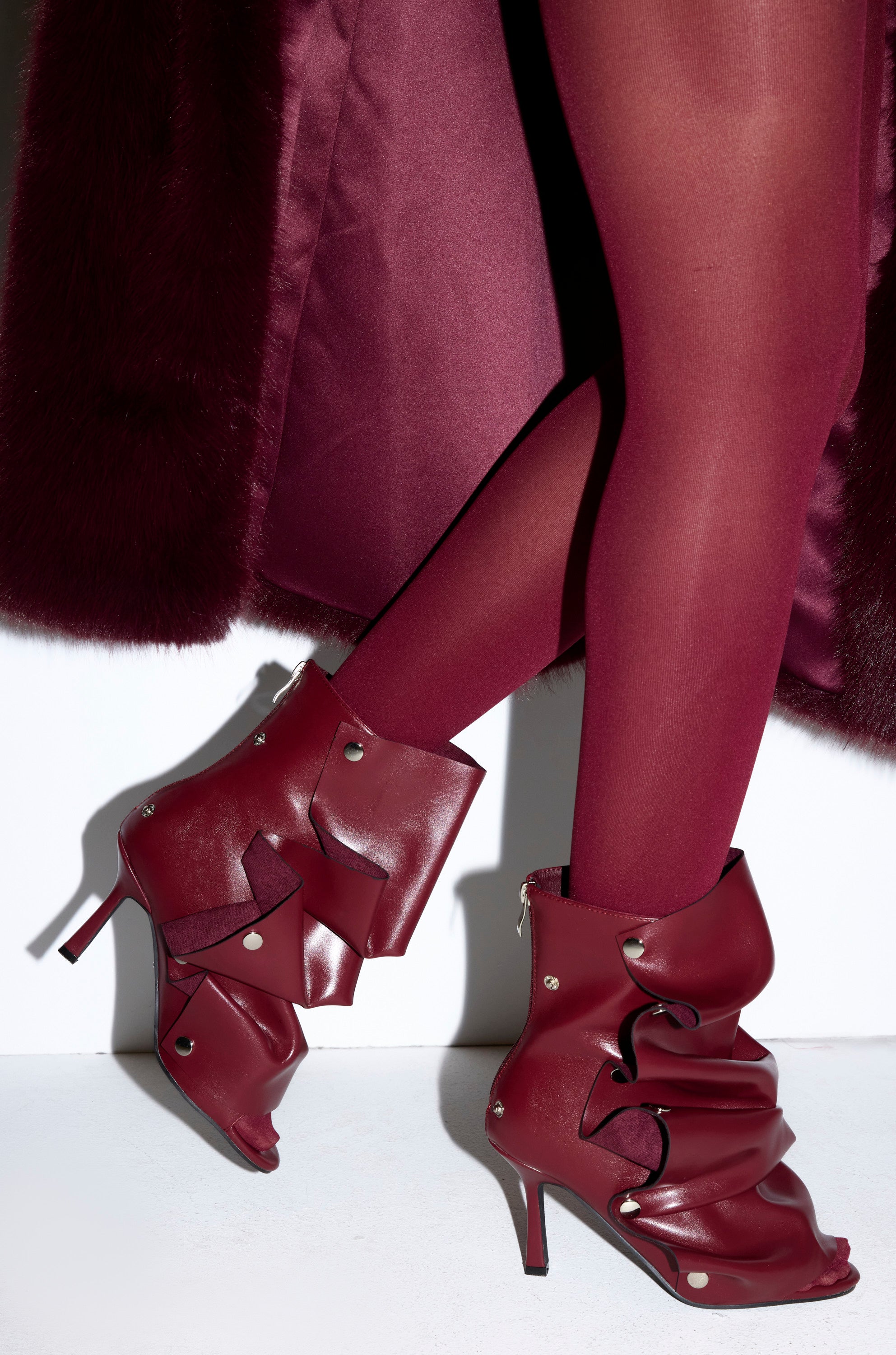 A woman stands against a white background in sheer burgundy tights, a matching coat, and the AZALEA WANG BIRDSONG BURGUNDY SNAP OPEN TOE STILETO BOOTIE featuring ruffled, layered leather and silver rivet accents.