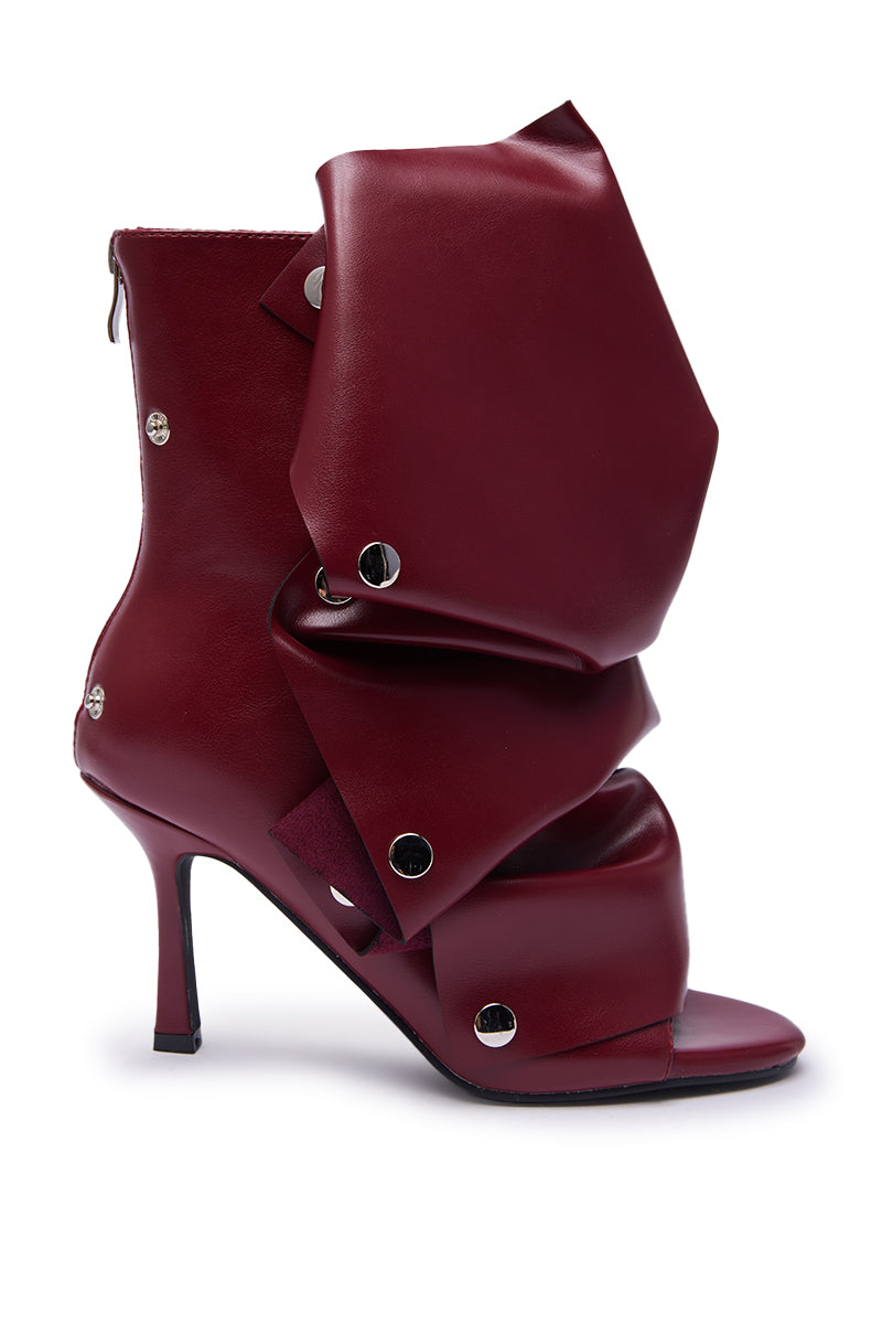 The AZALEA WANG BIRDSONG BURGUNDY SNAP OPEN TOE STILETTO BOOTIE features an open toe design, bold oversized folded faux leather details, and metallic rivets for a sculptural, avant-garde look.
