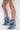 A person wearing a flowing white lace dress and the AZALEA WANG BIRD SONG DENIM OPEN TOE SNAP BOOTIE, featuring unique open toes, stiletto heels, and metal studs, stands on a white background.
