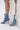 A person wears the AZALEA WANG BIRD SONG DENIM OPEN TOE SNAP BOOTIE, a light blue stiletto heel bootie with metal eyelets, on a white surface beside a flowing sheer white lace garment.