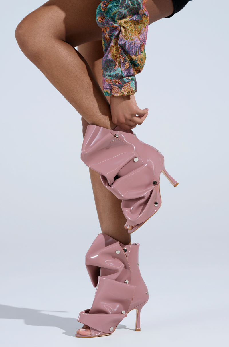 A person wearing the AZALEA WANG BIRD SONG SNAP OPEN TOE BOOTIE IN MAUVE, featuring a stylish ruched design and decorative buttons. Crafted from faux patent leather, its stiletto heel complements a colorful sleeve against a light grey background.
