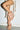 A person stands with one leg crossed, wearing draped, mocha AZALEA WANG BIRDSONG SNAP OPEN-TOE BOOTIES and a garment adorned with long, thick, white rope-like fringes.
