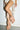 Wearing red nail polish and the AZALEA WANG BIRDSONG Snap Open-Toe Bootie in Mocha, a person stands against a plain wall in an outfit featuring thick white rope-like strands hanging down.