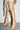 A person indoors models a cream skirt with long fringe, revealing bare legs and AZALEA WANG BIRDSONG SNAP OPEN-TOE BOOTIE IN MOCHA—light pink stilettos featuring decorative studs.