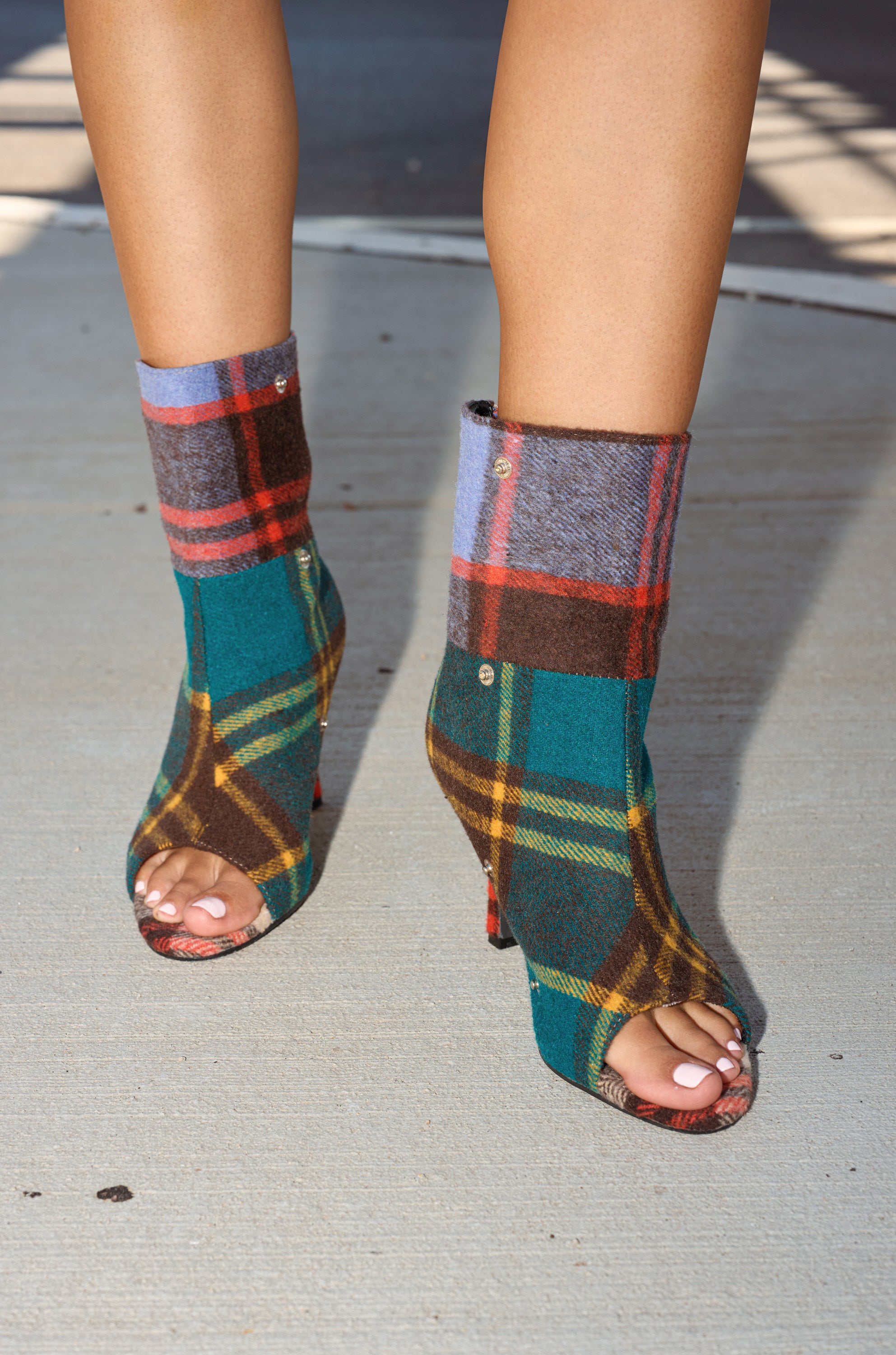 A person stands on a concrete surface wearing the AZALEA WANG BIRD SONG PLAID MULTI OPEN TOE SNAP BOOTIE, featuring a vibrant green, yellow, brown, and blue plaid pattern.