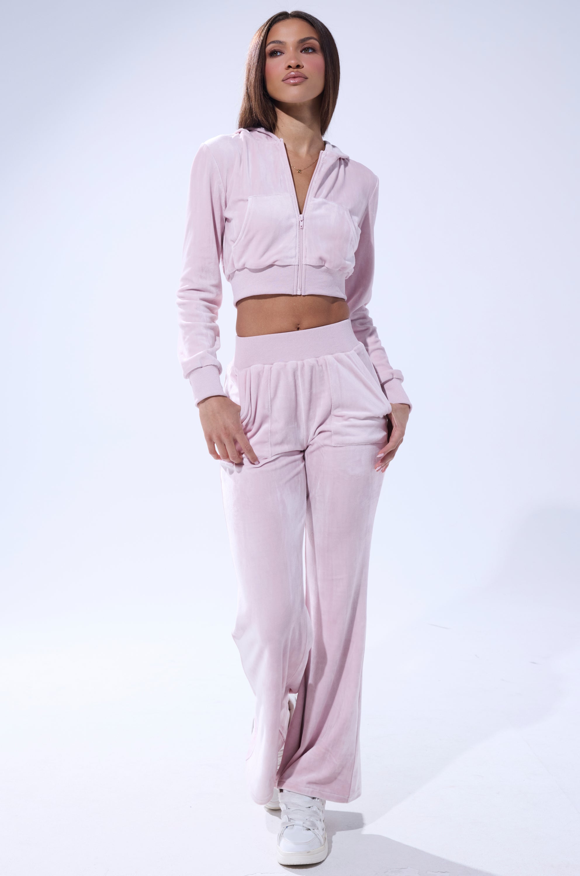 A woman wears the Taylor Straight Leg Jogger in Pink, featuring a cropped zip-up hoodie and high-rise wide-leg velour pants with white sneakers, set against a plain white background. 7
