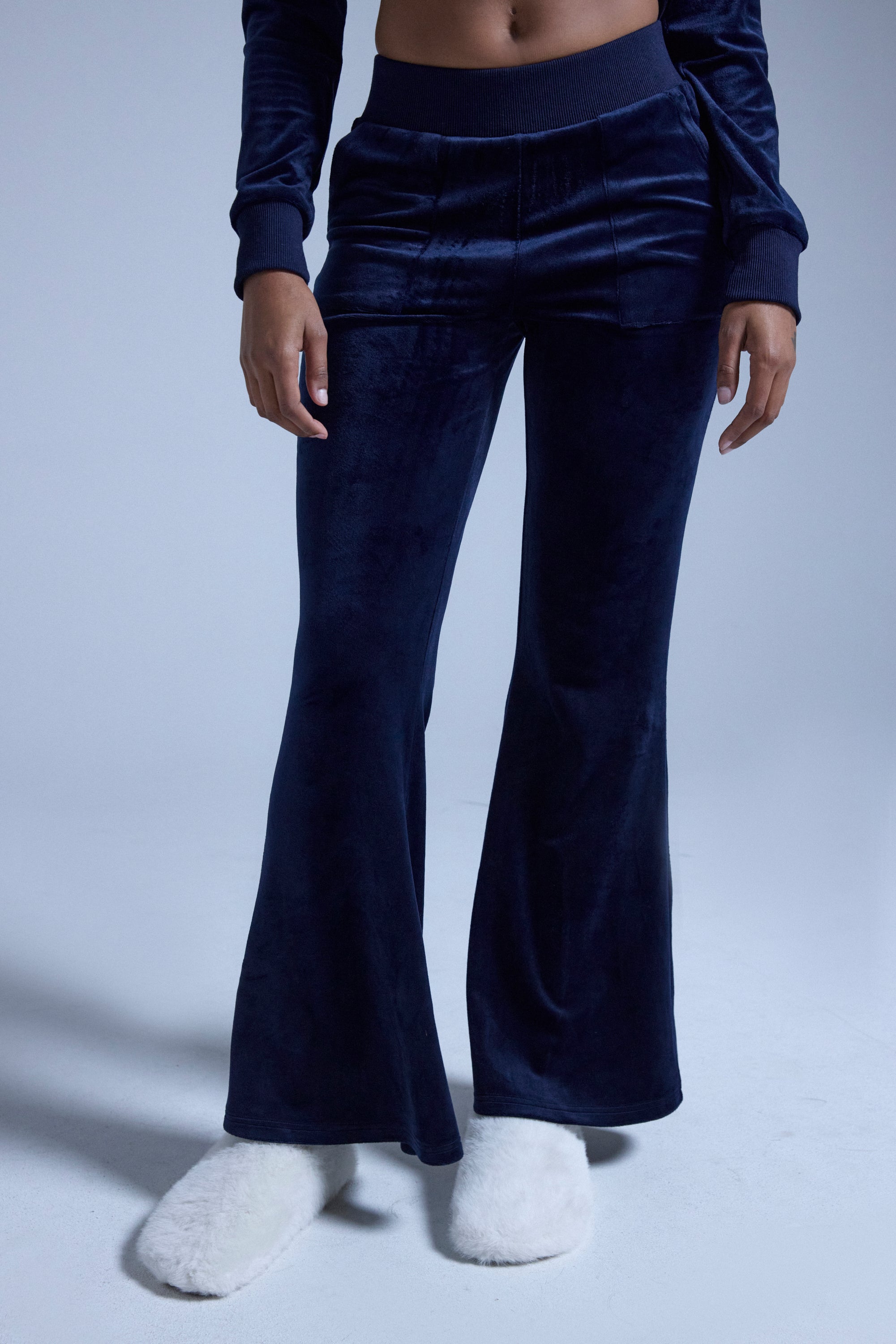 A person wearing the TAYLOR STRAIGHT LEG JOGGER IN NAVY with a matching top and white fuzzy slippers stands against a plain light blue background, with their upper body partially out of frame. 1
