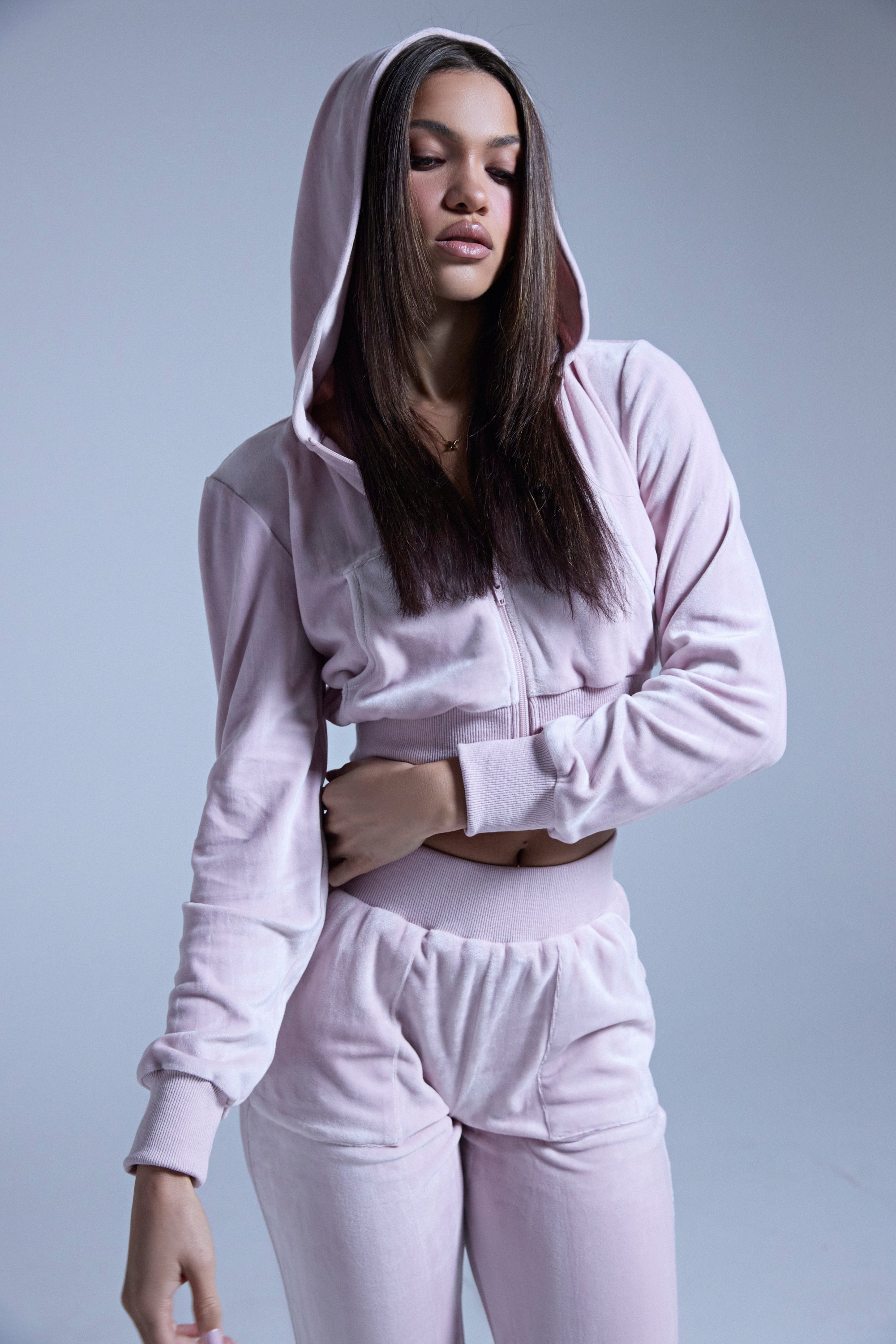 A woman with straight brown hair wears the TAYLOR VELOUR FITTED ZIP UP IN PINK—a light pink cropped hoodie and matching pants—standing against a plain background with her hood up and eyes closed. 0