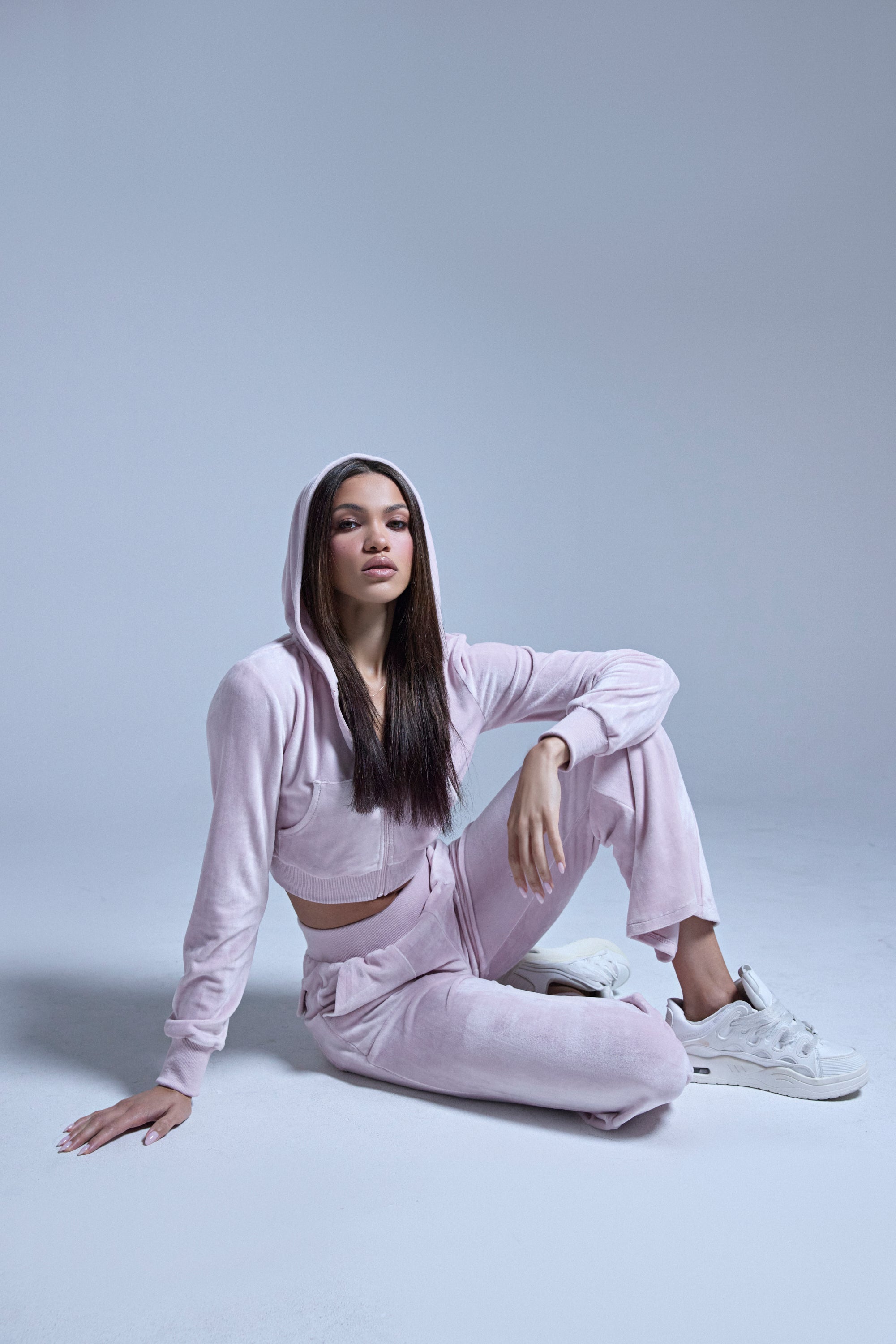 Wearing the TAYLOR VELOUR FITTED ZIP UP IN PINK and white sneakers, a woman sits on the floor against a plain light gray background, one knee raised and her hand resting on the ground. 4