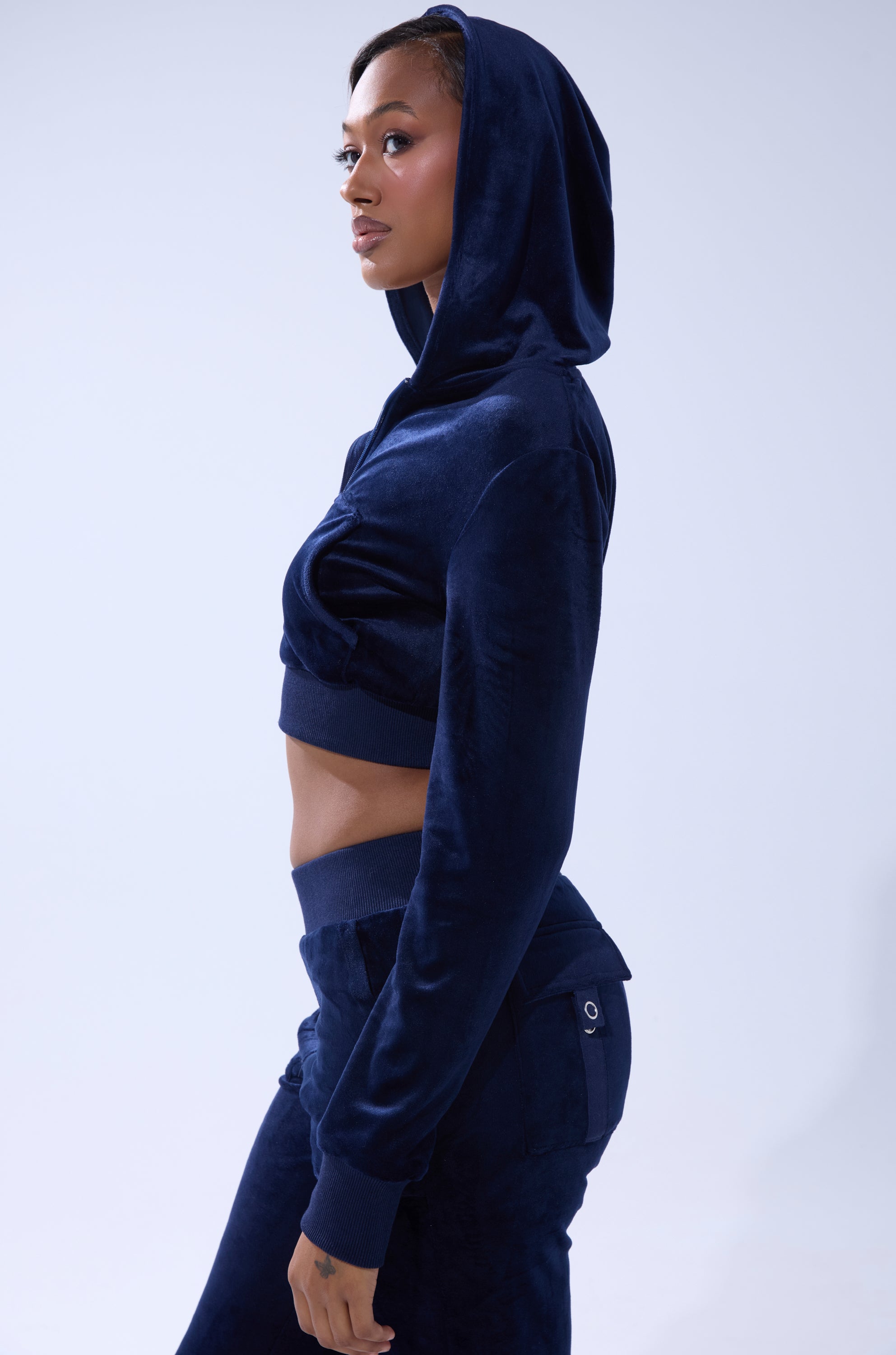 A woman wears the TAYLOR VELOUR FITTED ZIP UP IN NAVY—a cropped hoodie and pants set—standing in profile against a light background with the hood up, showcasing effortless loungewear style. 2