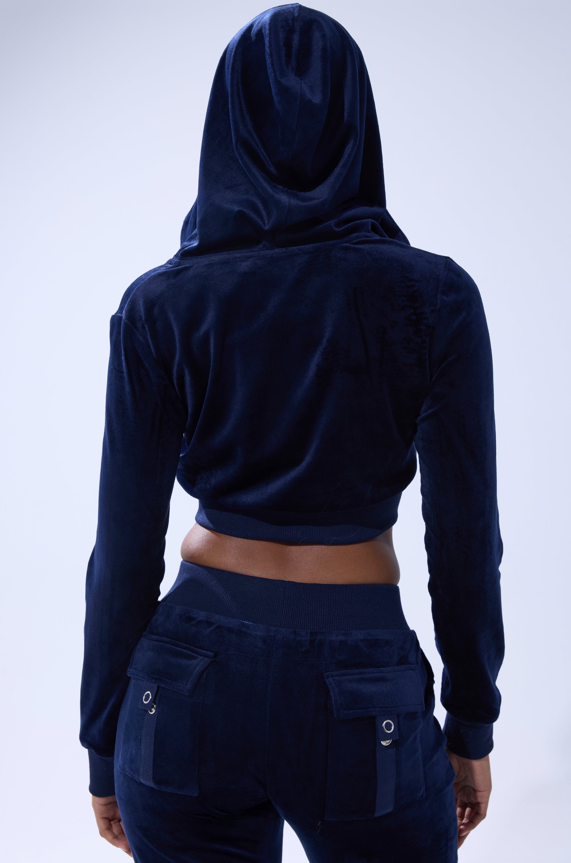 A person is seen from the back wearing the TAYLOR VELOUR FITTED ZIP UP IN NAVY with the hood up and matching flap-pocket loungewear bottoms, against a soft light blue background. 3