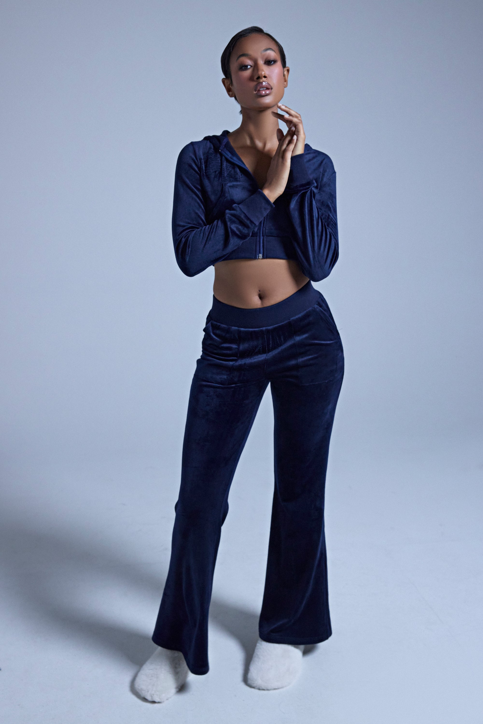 A woman poses in a studio wearing the TAYLOR VELOUR FITTED ZIP UP IN NAVY, styled with matching cropped pants and white fuzzy slippers, her hands near her chin against a plain light background. 4