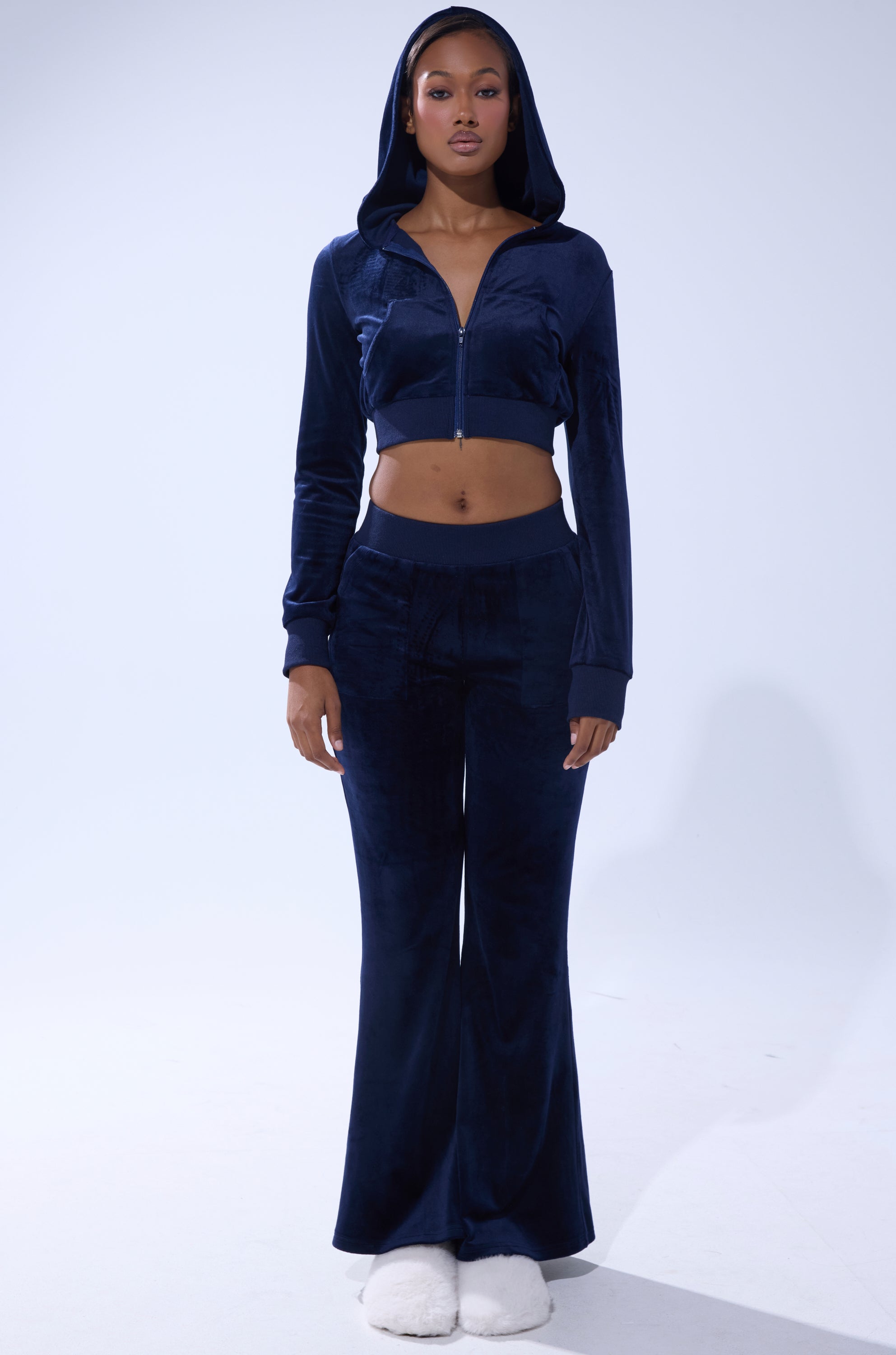 A person stands facing forward in the TAYLOR VELOUR FITTED ZIP UP IN NAVY, paired with matching high-waisted flared pants and white slippers, against a plain light background. 5