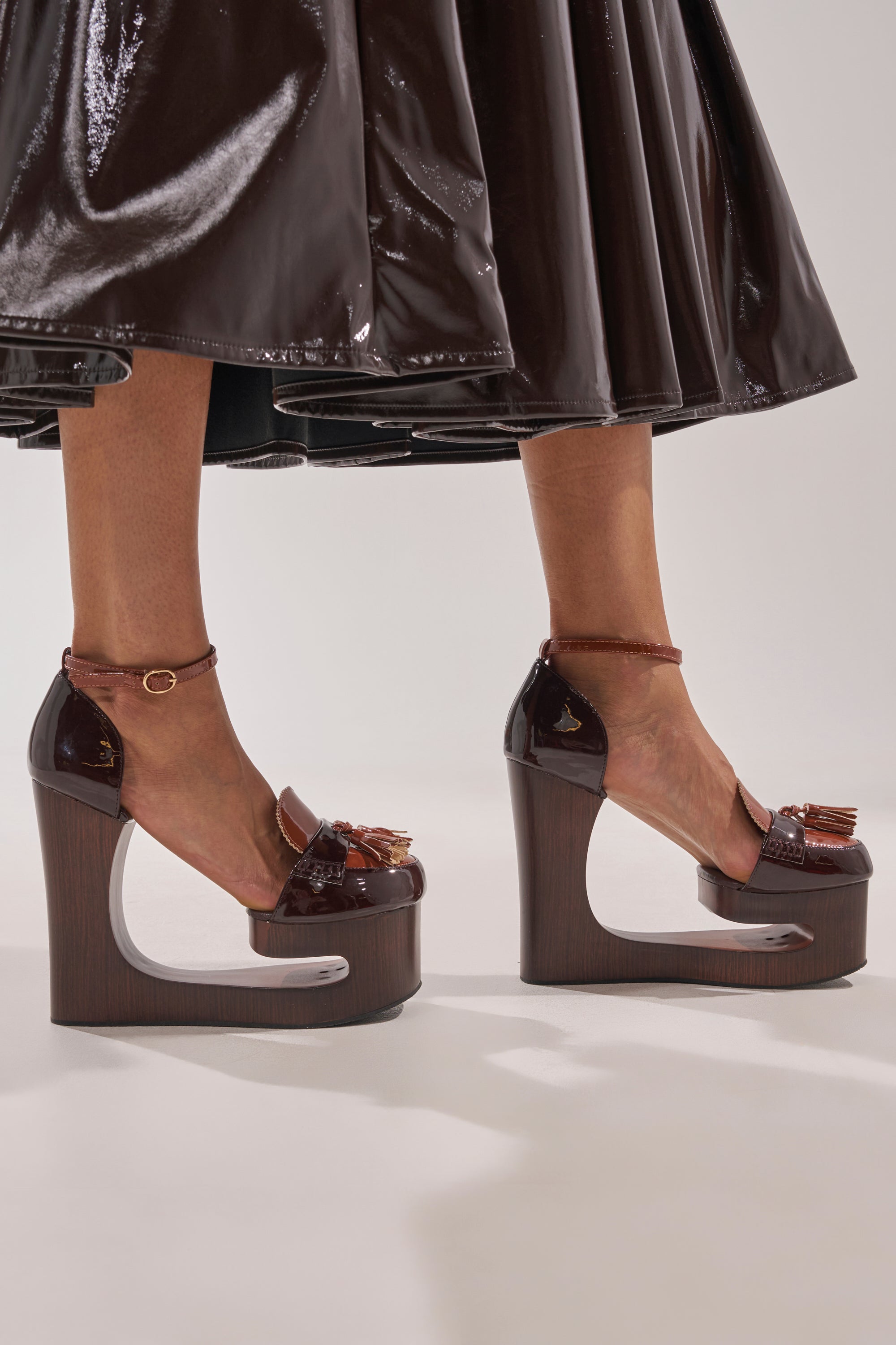 A person wearing the AKIRA GOLD BIXBY BROWN EMPTY WEDGE PUMP, featuring shiny dark brown faux leather with sculptural cutout heels and ankle straps, paired with a glossy dark brown skirt.