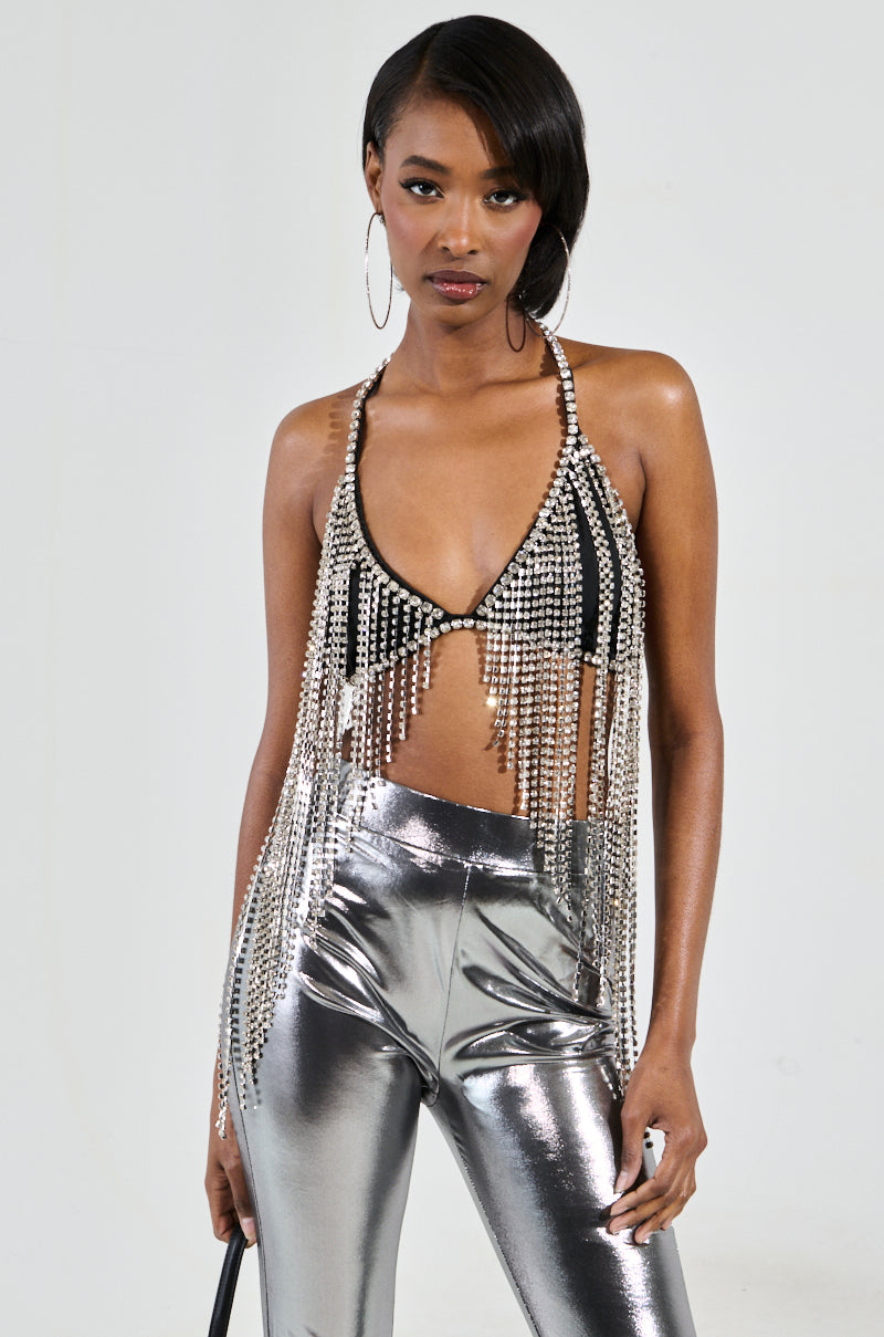 A woman stands against a plain background wearing a party-perfect piece: the BRING THE FUN RHINESTONE FRINGE BRALETTE TOP, complemented by shiny silver pants. Her long, dark hair cascades gracefully as hoop earrings glint at her ears, with one hand resting elegantly at her side.