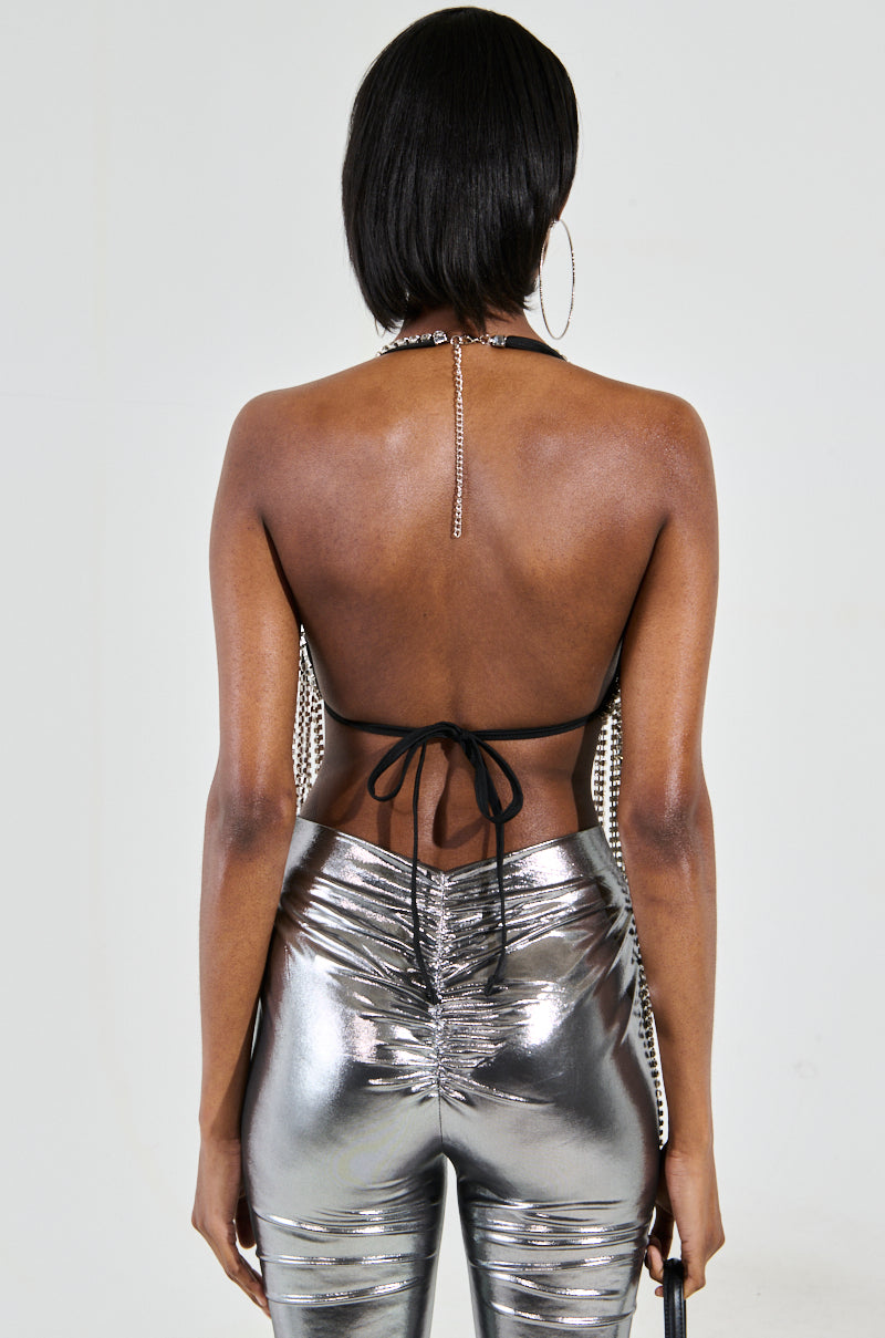 The person with short black hair is styled in the party-ready 'Bring the Fun Rhinestone Fringe Bralette Top', featuring a metallic design with sparkling silver trim. They pair this standout top, tied with black strings, with shiny silver pants and large hoop earrings, standing against a simple light backdrop.
