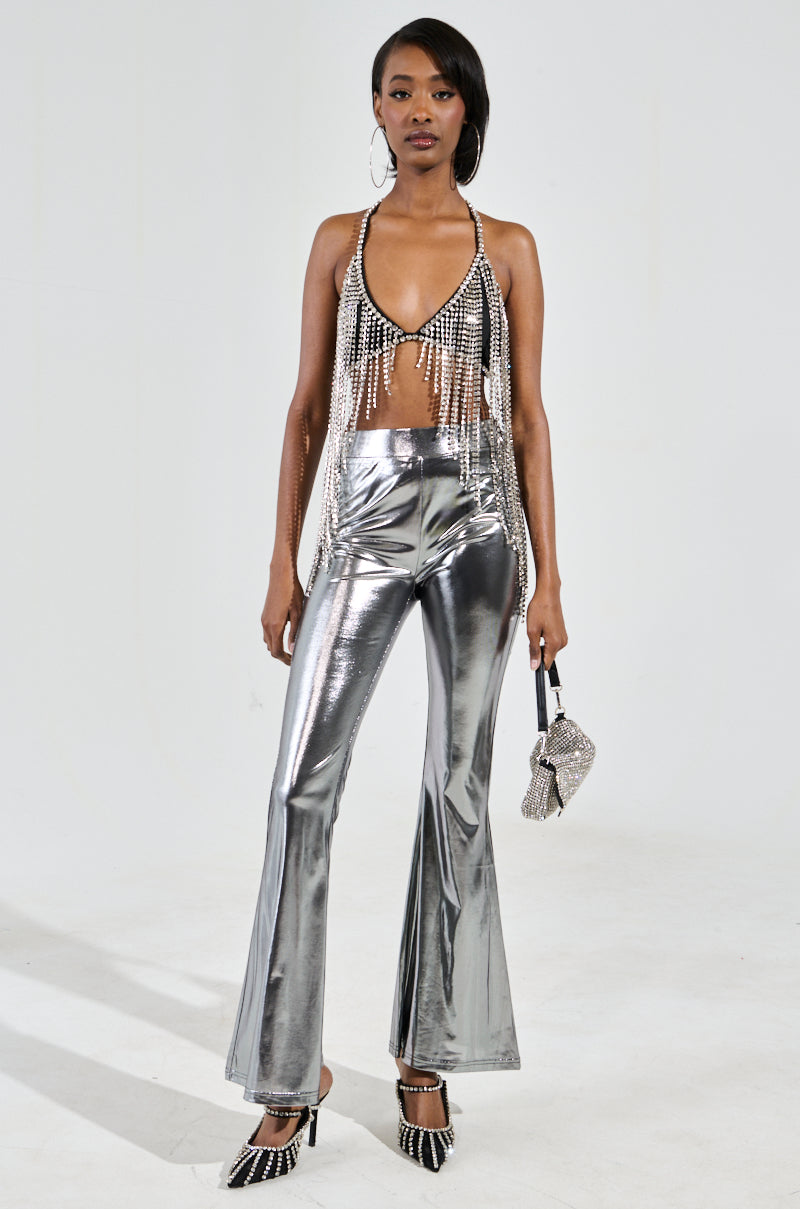 A woman captivates in a party-ready ensemble, wearing the BRING THE FUN RHINESTONE FRINGE BRALETTE TOP paired with metallic silver pants. Against a plain backdrop, she completes her look with sparkling silver trim on her shoes and carries a small silver handbag, emanating glamorous confidence.