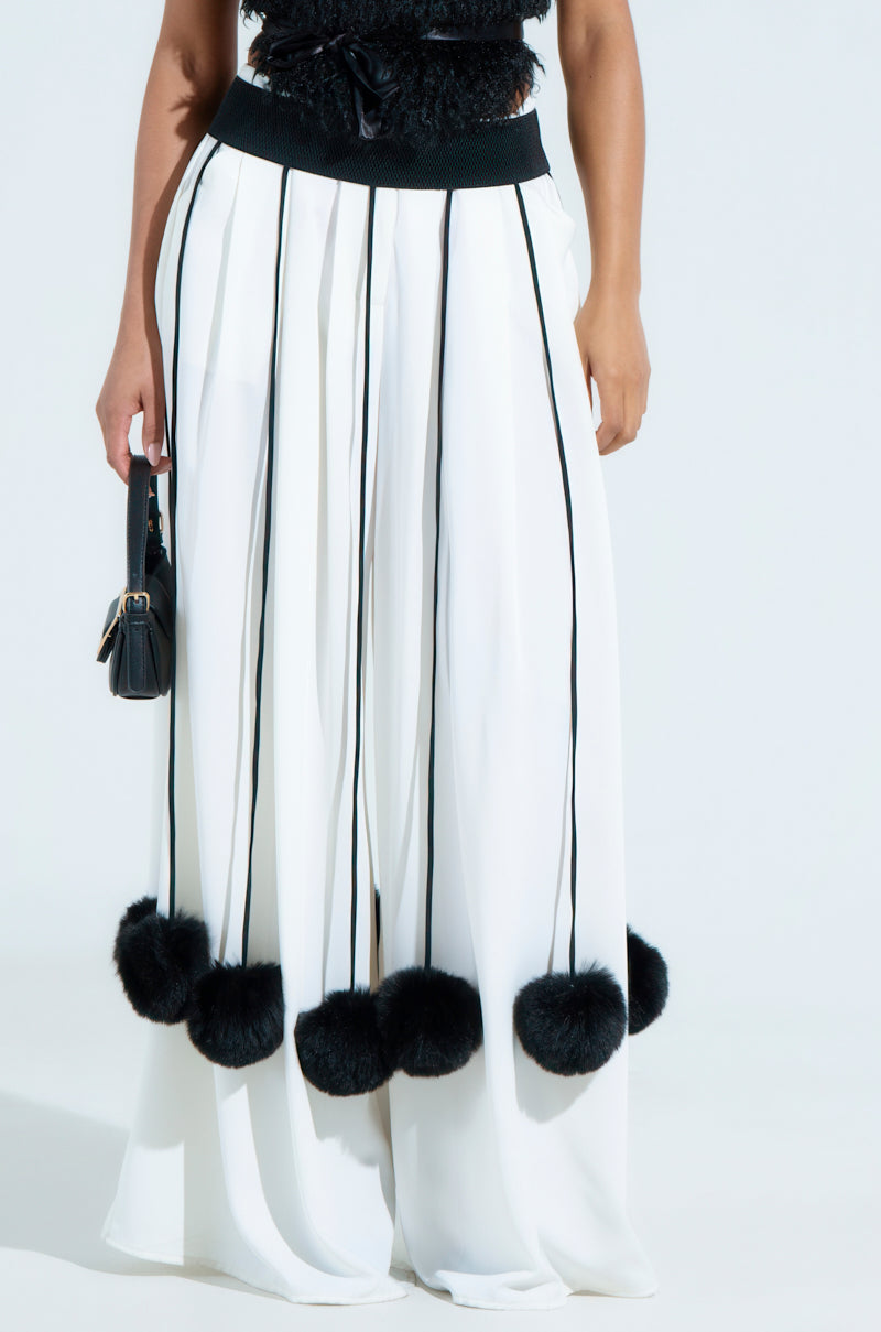 A person wearing a white pleated skirt featuring black vertical stripes and adorned with the SHAKE YA POM POMS FUR BELT carries a small black handbag. The upper outfit shows a rich black texture.