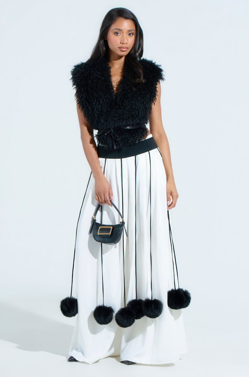 A woman poses against a plain backdrop in a black, fuzzy sleeveless SHAKE YA POM POMS FUR BELT top paired with a long white fringed skirt featuring pom-poms. She accessorizes with a small black handbag while facing the camera.