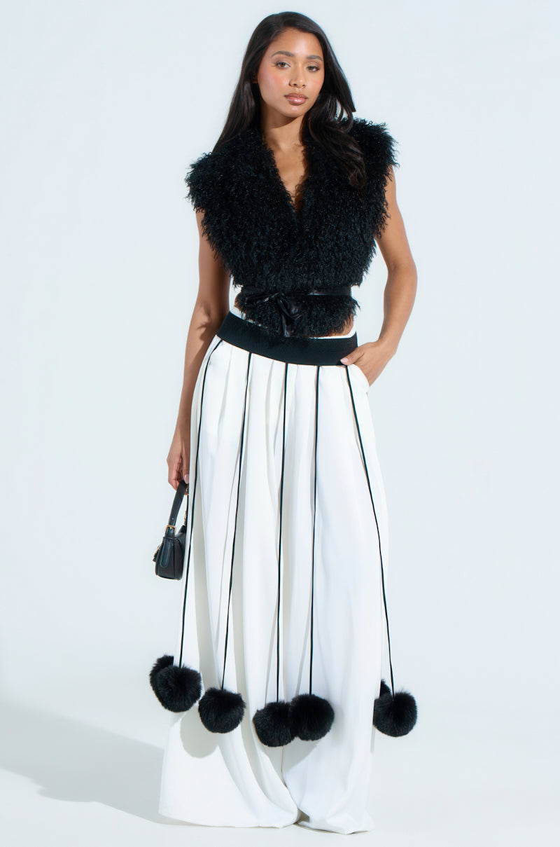 A woman exudes confidence in a sleeveless black fur top paired with the SHAKE YA POM POMS FUR BELT. Her long white skirt has vertical black stripes and faux fur pom-poms. She holds a small black handbag against a plain white backdrop.