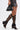 A person wears a short dark gray skirt, holds a small black handbag with metallic details, and sports AZALEA WANG BLACKBIRD BLACK PU STILETTO GLADIATOR SANDALS. The background is plain white.