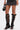 Someone is wearing a short dark skirt and AZALEA WANG BLACKBIRD BLACK PU STILETTO GLADIATOR SANDALS with crisscross straps, open toes, and stiletto heels. They're holding a black handbag against a plain white background.