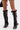 A person wears the AZALEA WANG BLACKBIRD BLACK PU STILETTO GLADIATOR SANDAL—unique, black, knee-high with bold crisscross straps and open toes—paired with a short skirt. Only the legs are shown against a plain background.
