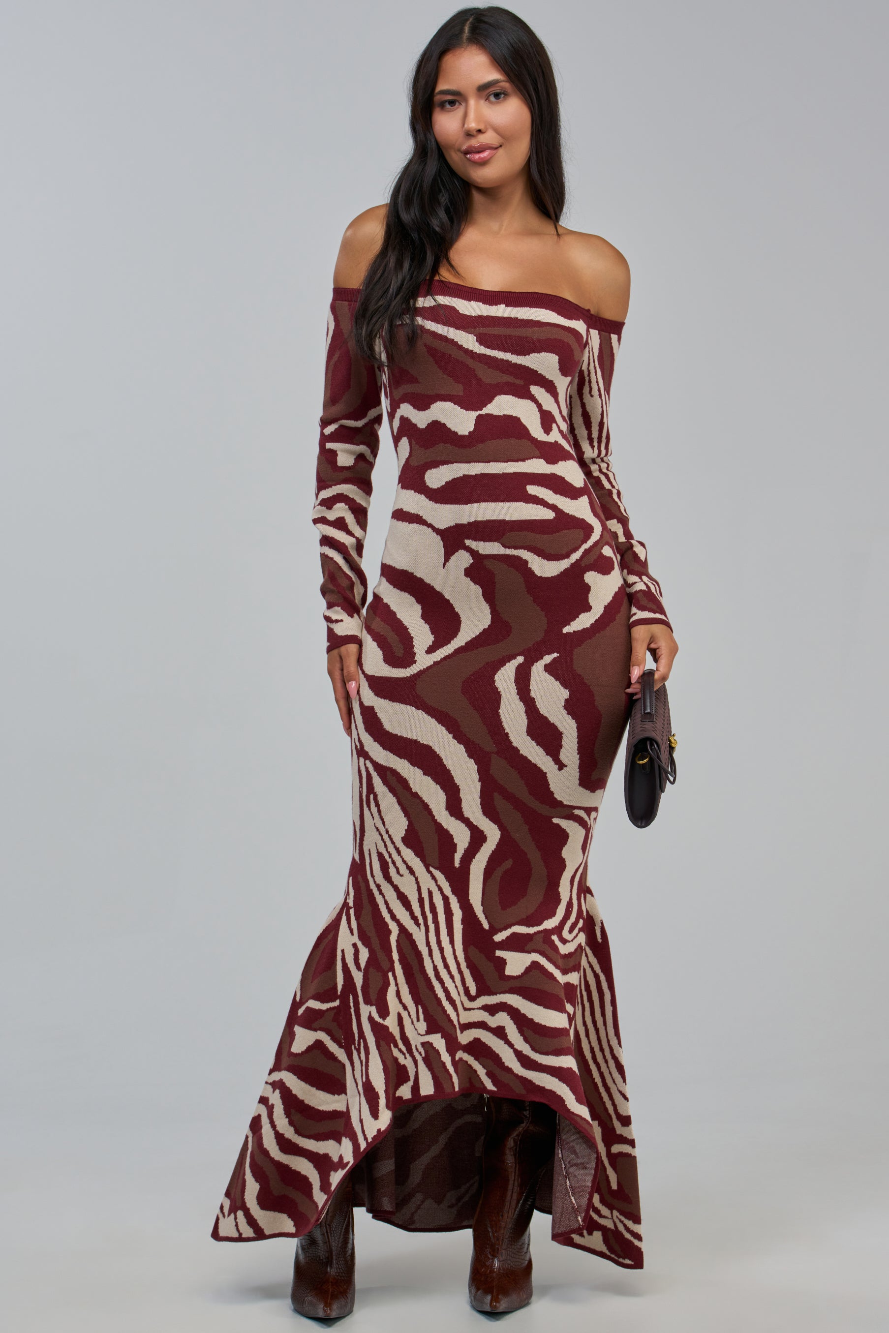 A woman wearing the KALLIE OFF THE SHOULDER LONG SLEEVE SWEATER DRESS, which features a maroon and beige abstract zebra print and long sleeves, holds a black clutch while standing against a plain gray background.