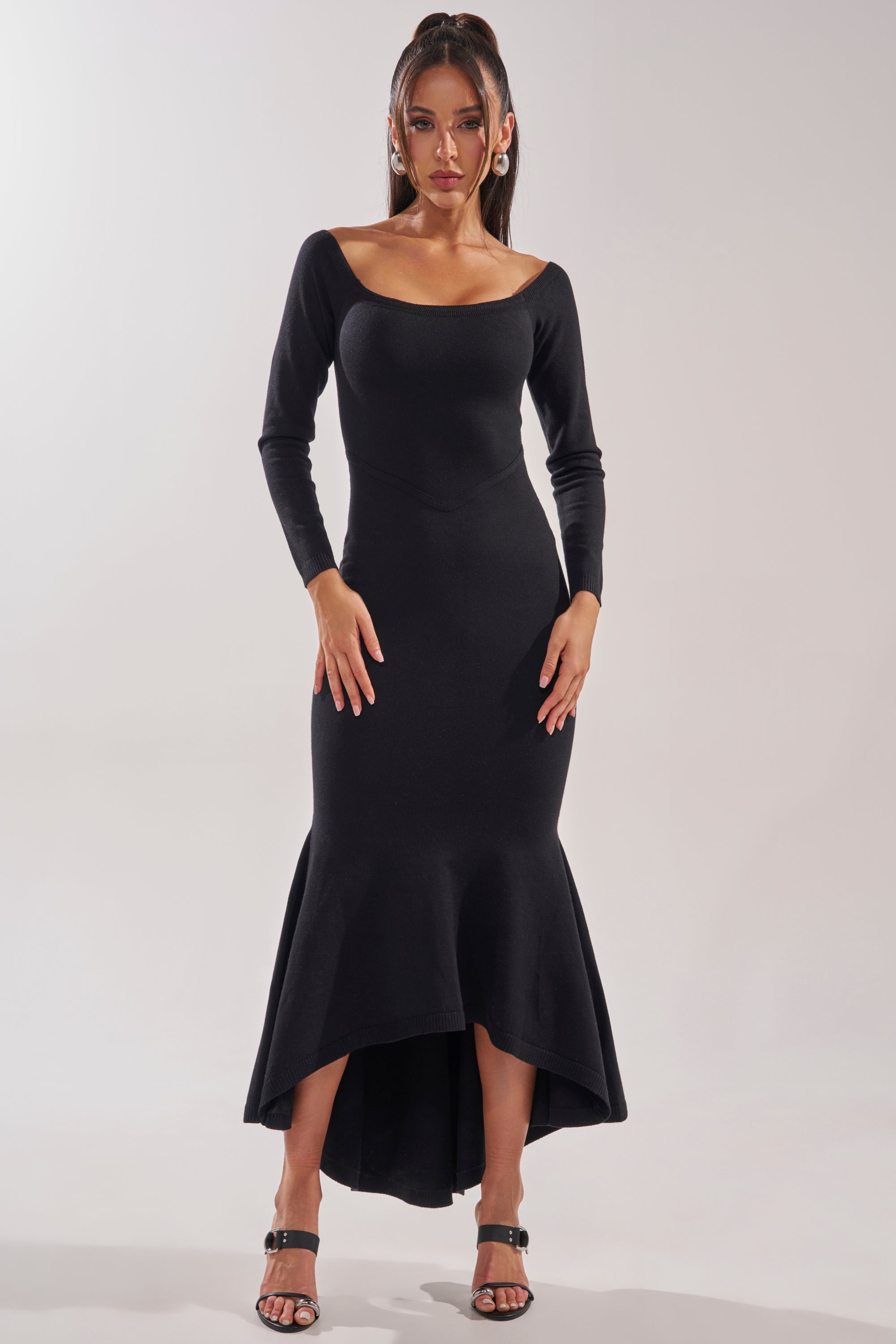 A woman with long hair and large hoop earrings poses against a plain background in the KALLIE OFF SHOULDER LONG SLEEVE SWEATER MAXI DRESS, featuring a mermaid silhouette, flared high-low hem, and black heeled sandals.