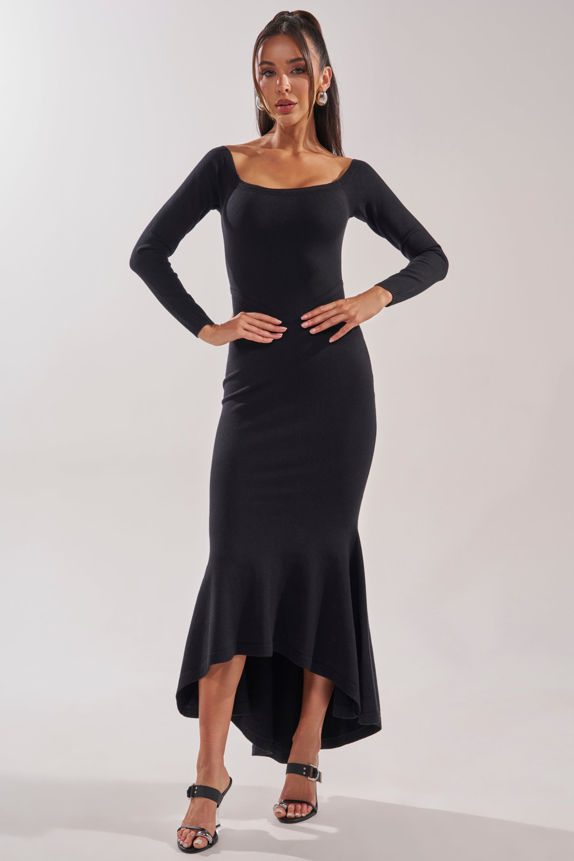 A woman models the KALLIE OFF SHOULDER LONG SLEEVE SWEATER MAXI DRESS, featuring a fitted black knit, long sleeves, and mermaid silhouette, styled with black heeled sandals against a plain light backdrop. 1