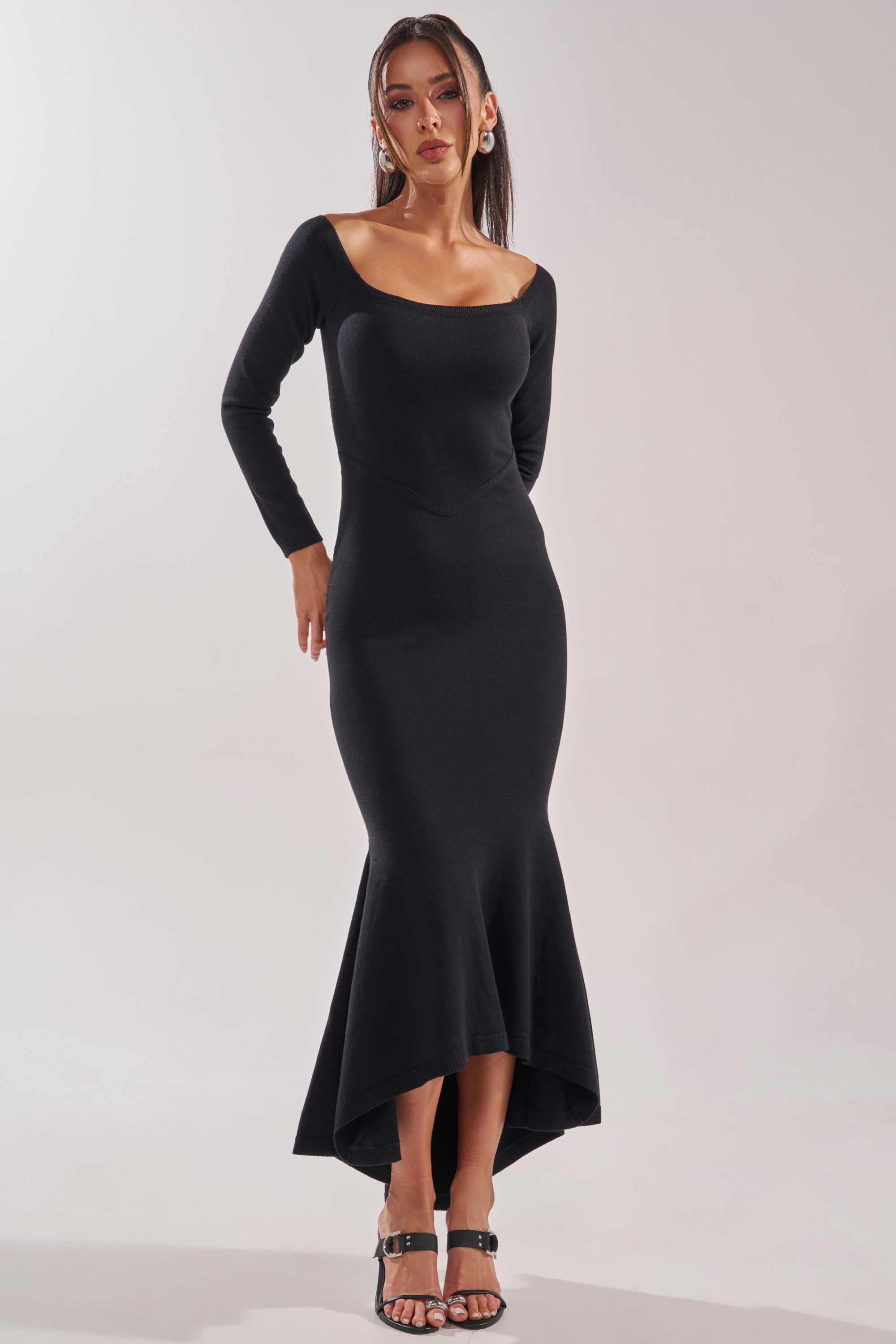 A woman poses against a plain light background wearing the KALLIE OFF SHOULDER LONG SLEEVE SWEATER MAXI DRESS in black, featuring a fitted mermaid silhouette, long sleeves, off-shoulder neckline, and high-low hem with black heeled sandals. 2