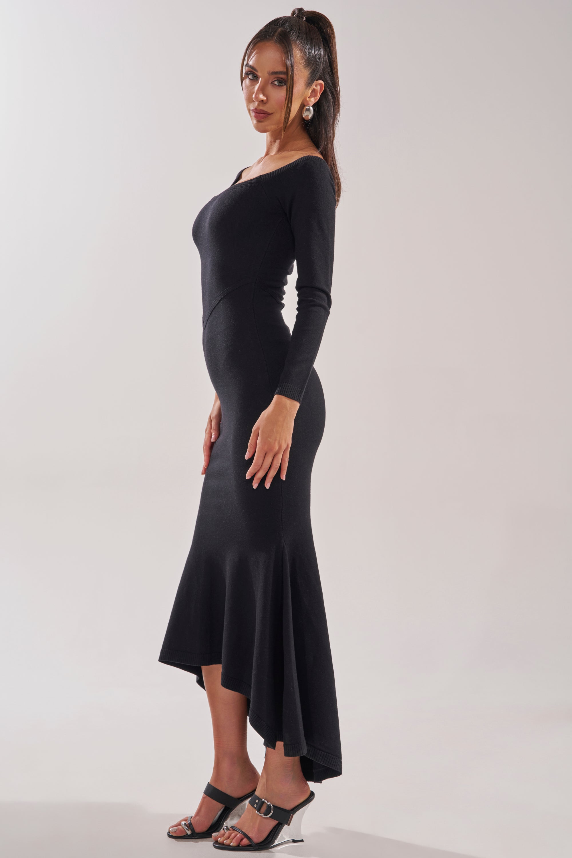 A woman stands sideways in the KALLIE OFF SHOULDER LONG SLEEVE SWEATER MAXI DRESS, featuring a fitted black knit design and asymmetrical flared hem. She pairs it with black heeled sandals, long hair in a high ponytail, against a plain light background. 3
